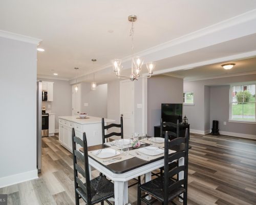 Open-concept dining area with a rectangular table set for four, black chairs, and a bright kitchen island in a light-gray home interior.
