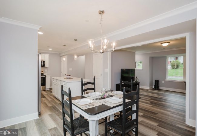 Open-concept dining area with a rectangular table set for four, black chairs, and a bright kitchen island in a light-gray home interior.