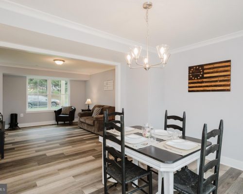 Open-concept dining area with a white table, black chairs, placemats, and a pendant chandelier; living room beyond.