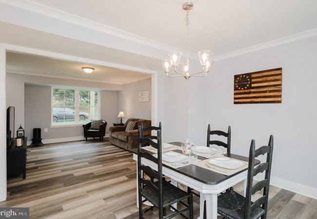Open-concept dining area with a white table, black chairs, placemats, and a pendant chandelier; living room beyond.