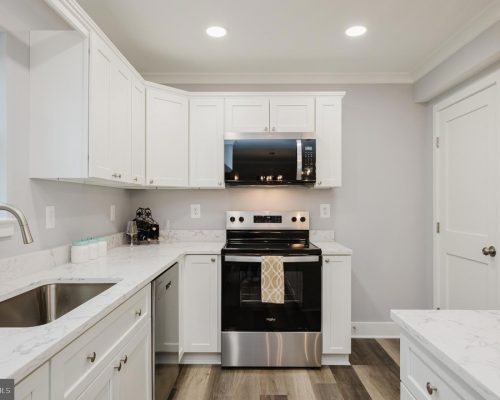 Modern white kitchen with marble countertops, stainless steel range and microwave, and a double sink on the left.