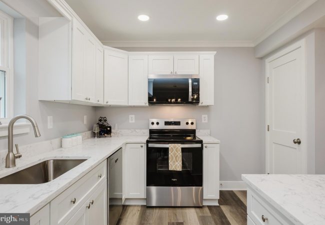 Modern white kitchen with marble countertops, stainless steel range and microwave, and a double sink on the left.