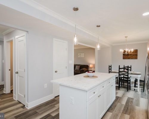 Open-concept kitchen with white island and marble countertop, pendant lights, and a dining area with black chairs in the background.
