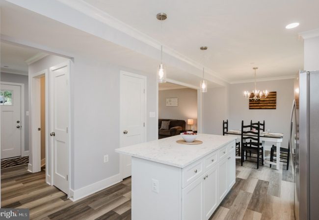 Open-concept kitchen with white island and marble countertop, pendant lights, and a dining area with black chairs in the background.