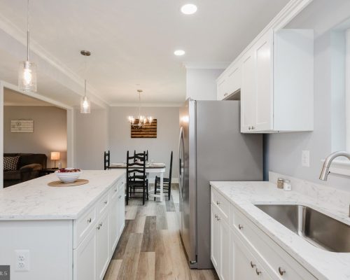 Bright modern kitchen with white cabinets, marble countertops, an island, and a stainless steel refrigerator; dining table visible in the back.