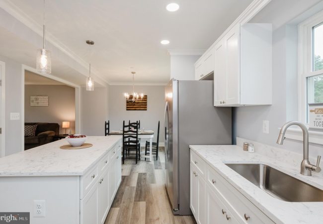 Bright modern kitchen with white cabinets, marble countertops, an island, and a stainless steel refrigerator; dining table visible in the back.