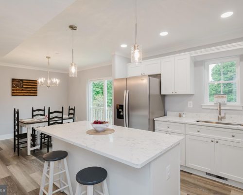 Bright modern kitchen with white cabinets, marble island, and stainless steel refrigerator; open dining area nearby with dark wooden chairs and a rectangular table.