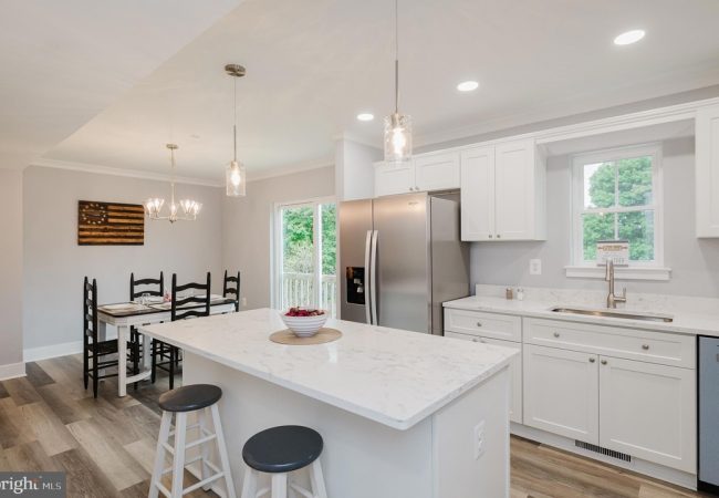 Bright modern kitchen with white cabinets, marble island, and stainless steel refrigerator; open dining area nearby with dark wooden chairs and a rectangular table.