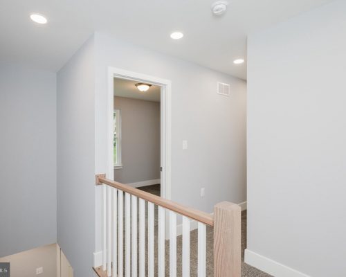 Bright upstairs hallway with gray walls, white trim, a wooden handrail and white balusters overlooking a staircase, and an open doorway to a room with a window.