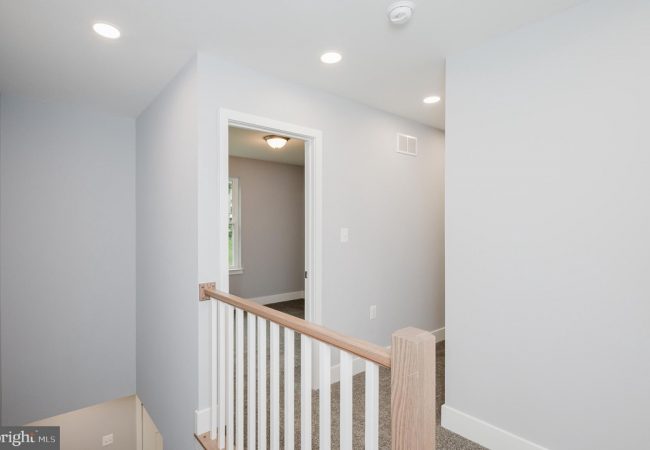 Bright upstairs hallway with gray walls, white trim, a wooden handrail and white balusters overlooking a staircase, and an open doorway to a room with a window.