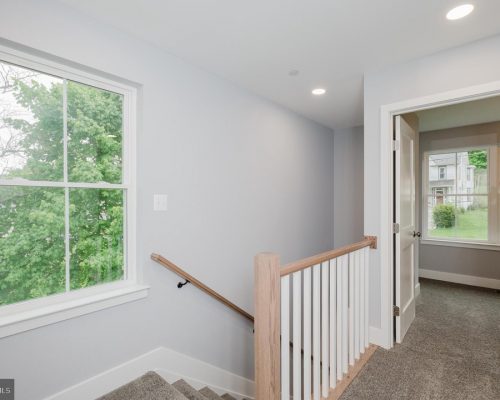 Interior stair landing with gray walls, carpeted stairs, and a wooden handrail leading down; bright window shows green trees outside and a doorway to an adjoining room.