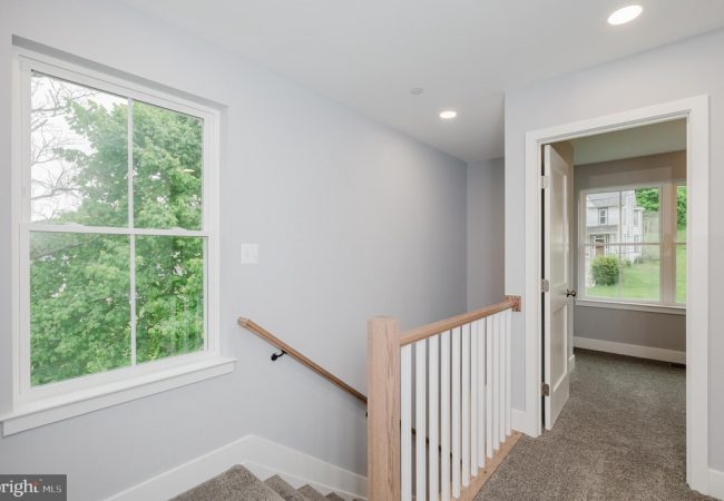 Interior stair landing with gray walls, carpeted stairs, and a wooden handrail leading down; bright window shows green trees outside and a doorway to an adjoining room.