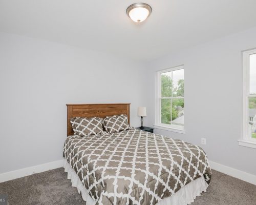 Bedroom with a wooden headboard queen bed, gray geometric duvet, two windows showing greenery, and a nightstand with a lamp on the right side.