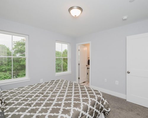 Bright bedroom with light gray walls, two large windows overlooking green trees, a bed with a gray-and-white patterned duvet, and a doorway to an en-suite bathroom.