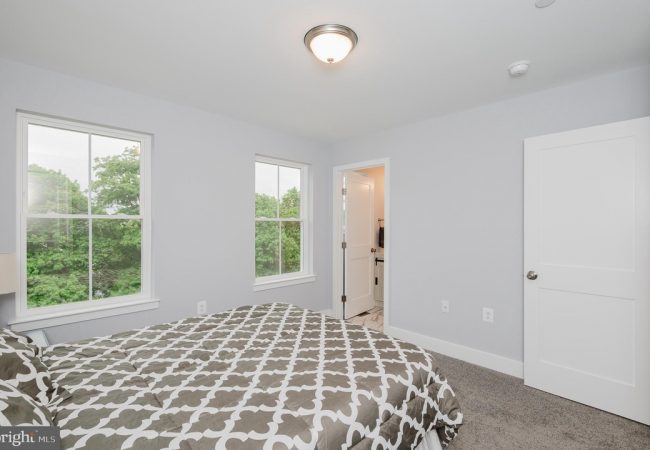 Bright bedroom with light gray walls, two large windows overlooking green trees, a bed with a gray-and-white patterned duvet, and a doorway to an en-suite bathroom.