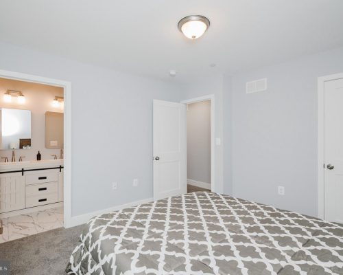Bedroom with a gray-patterned comforter, light gray walls, and an open doorway to a white-turnished bathroom with double sinks and mirrors on marble floor.
