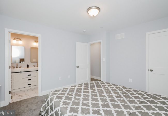 Bedroom with a gray-patterned comforter, light gray walls, and an open doorway to a white-turnished bathroom with double sinks and mirrors on marble floor.