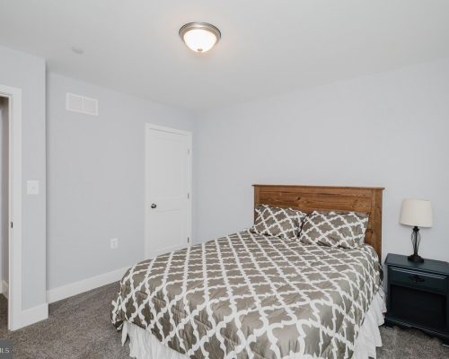 Bedroom with a wooden headboard bed, gray geometric bedding, two pillows, a dark nightstand with lamp, and pale gray walls.