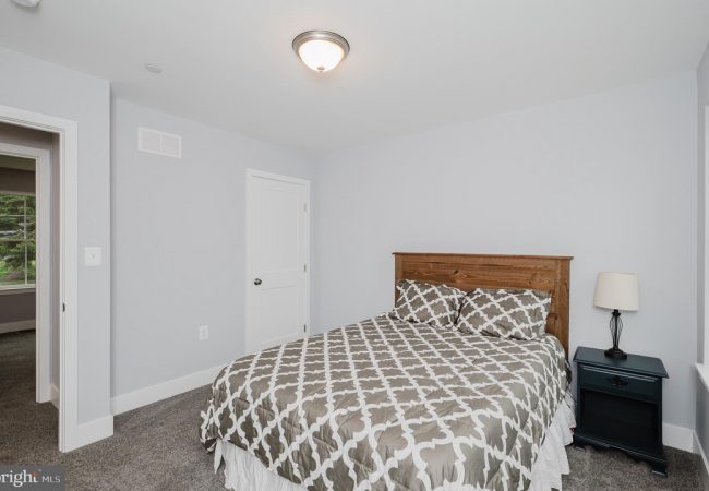Bedroom with a wooden headboard bed, gray geometric bedding, two pillows, a dark nightstand with lamp, and pale gray walls.