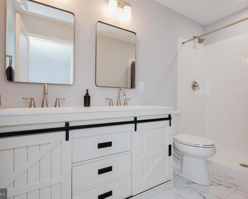 Double-sink bathroom vanity with white cabinets, black hardware, and sliding barn-door drawers, in a light gray room with two rectangular mirrors and brushed-nickel faucets.
