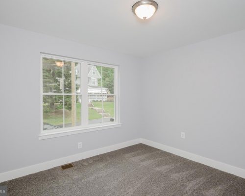 Empty bedroom with light gray walls, beige carpet, and a central ceiling light fixture; window shows a suburban street outside.