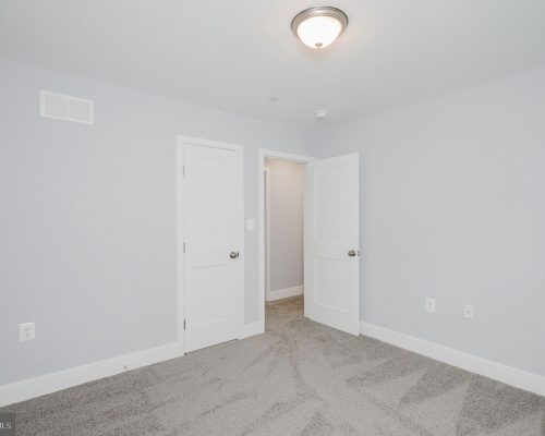 Empty bedroom with pale gray walls, white doors, and light gray carpet; ceiling light centered above.