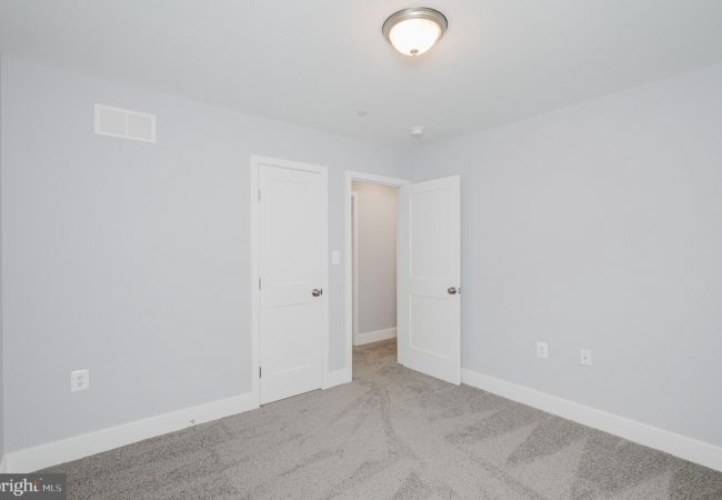 Empty bedroom with pale gray walls, white doors, and light gray carpet; ceiling light centered above.