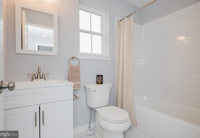 Bright white bathroom with a vanity, mirror, towel ring, toilet, and a bathtub with a beige curtain.
