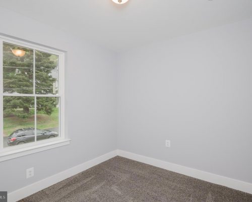 Empty light-gray room with white baseboards and a large window showing a parked car and greenery outside