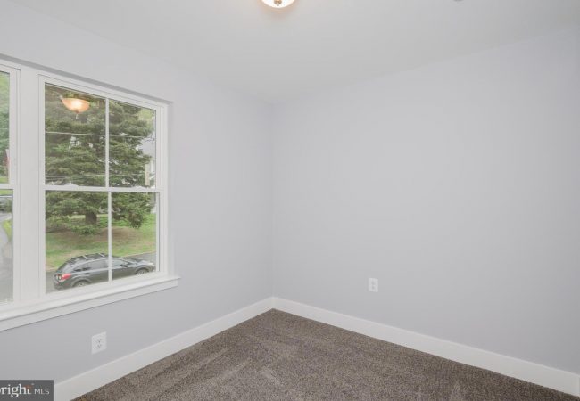 Empty light-gray room with white baseboards and a large window showing a parked car and greenery outside