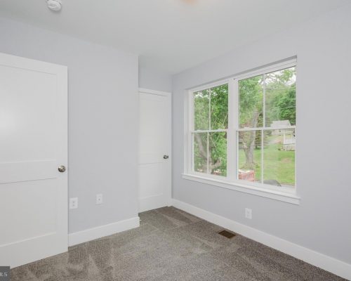 Empty bedroom with light gray walls, white doors, and a large window overlooking a green yard outside.