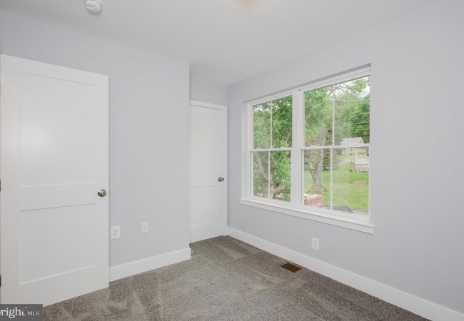 Empty bedroom with light gray walls, white doors, and a large window overlooking a green yard outside.