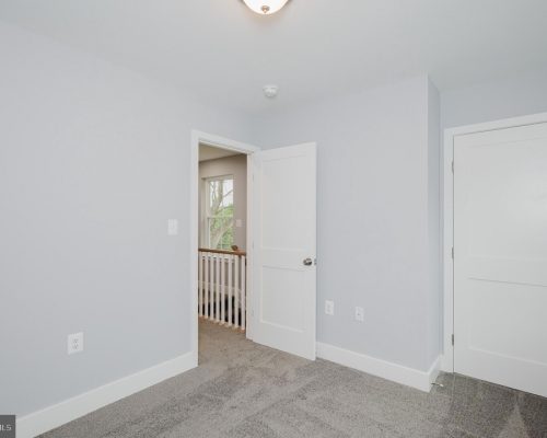 Empty light-blue bedroom with white doors, gray carpet, and baseboard trim; hallway visible beyond an open doorway.