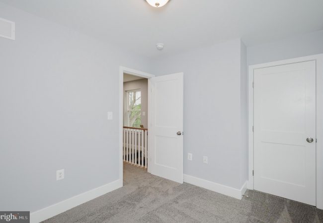 Empty light-blue bedroom with white doors, gray carpet, and baseboard trim; hallway visible beyond an open doorway.