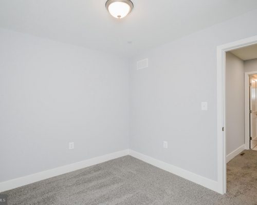 Empty small bedroom with light gray walls, white baseboards, carpet, ceiling light, and an open doorway to a bathroom.