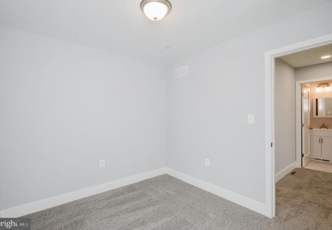 Empty small bedroom with light gray walls, white baseboards, carpet, ceiling light, and an open doorway to a bathroom.