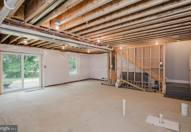 Unfinished basement with exposed joists, plastic-wrapped walls, and a sliding glass door to the outside area.