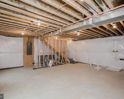 Unfinished basement with exposed ceiling joists, plastic walls, and construction tools scattered, including a ladder and stairs to an upper level for renovation work.