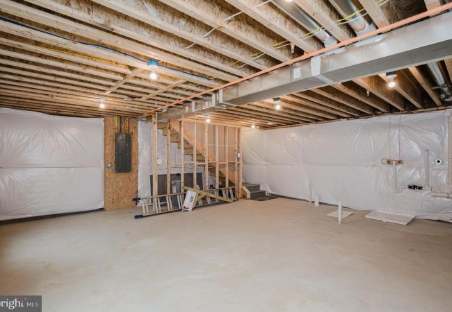 Unfinished basement with exposed ceiling joists, plastic walls, and construction tools scattered, including a ladder and stairs to an upper level for renovation work.