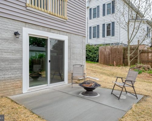 Backyard patio with sliding glass door, two metal chairs, and a round fire pit on a concrete slab.