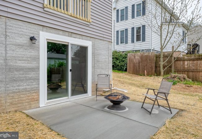 Backyard patio with sliding glass door, two metal chairs, and a round fire pit on a concrete slab.