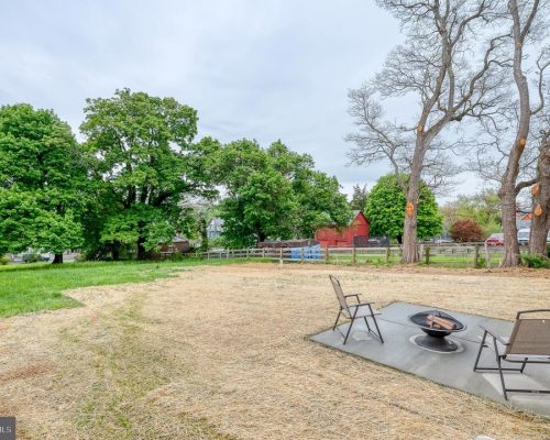 Backyard with a circular fire pit and three chairs on a concrete pad, straw-covered ground, and green trees in the background.