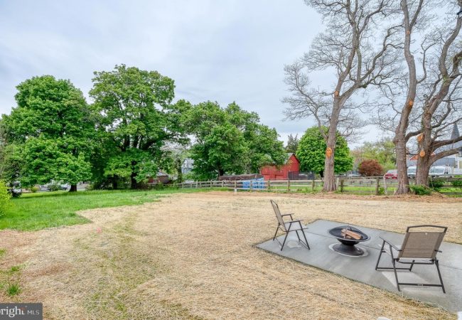 Backyard with a circular fire pit and three chairs on a concrete pad, straw-covered ground, and green trees in the background.