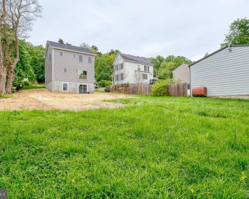 Wide view of a green backyard with two-story gray houses, a wooden fence, and a white shed in a suburban neighborhood.