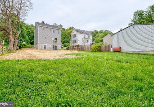 Wide view of a green backyard with two-story gray houses, a wooden fence, and a white shed in a suburban neighborhood.