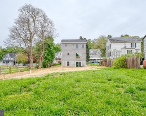 Backyard view with a three-story gray house in the center, a large bare tree on the left, and a wooden fence along the right edge; green lawn in the foreground.