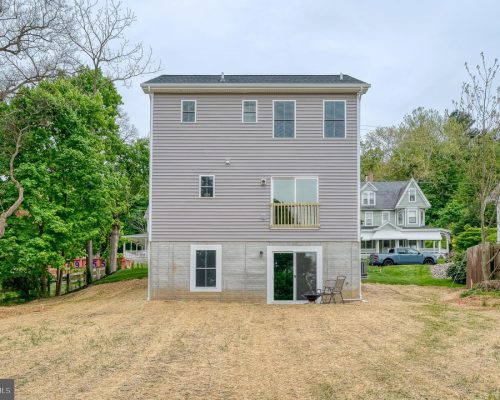 Three-story gray-siding house with a concrete basement level, back yard, sliding glass door, and a small balcony above the ground floor patio.