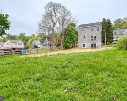 Green grassy lot in a residential area with a gray house under construction in the background and trees nearby as neighbors look on.