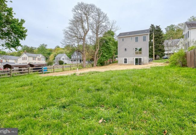 Green grassy lot in a residential area with a gray house under construction in the background and trees nearby as neighbors look on.