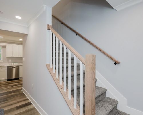 Staircase with light wood handrail and white balusters beside a hallway leading to a white kitchen with stainless appliances.